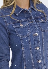 Denim jacket in blue with silver-toned studs. Features classic collar, two chest pockets, and button closures along the front.