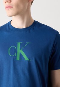 Plava pamučna majica s grafičkim logotipom u zelenoj boji koji prikazuje "CK" s natpisom "Calvin Klein" ispod. Kratki rukavi i dizajn s okruglim izrezom.