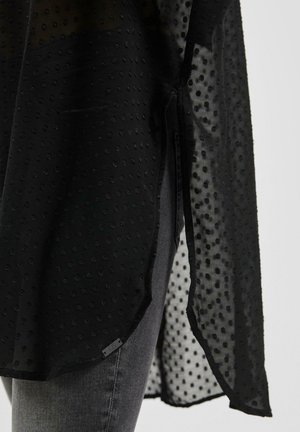 Durchsichtige schwarze Bluse mit einem strukturierten Punktmuster, ausgestattet mit seitlichen Schlitzen und einem geschwungenen Saum. Kombiniert mit grauen Jeans.