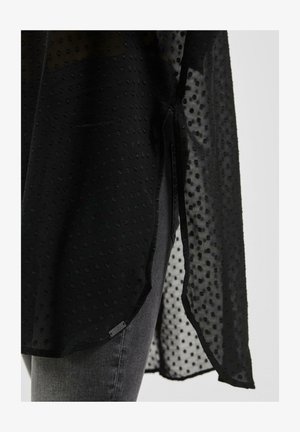 Permatomas juodas sijonas su tekstūruota taškų raštu, su šoninėmis plyšėmis ir kreipiančiu apdaila. Sukomplektuotas su pilkais džinsais.