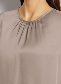 Beige Bluse mit gerafftem Ausschnitt, aus glattem Stoff gefertigt. Zeigt eine weiche, fließende Textur ohne sichtbare Muster oder Hardware.