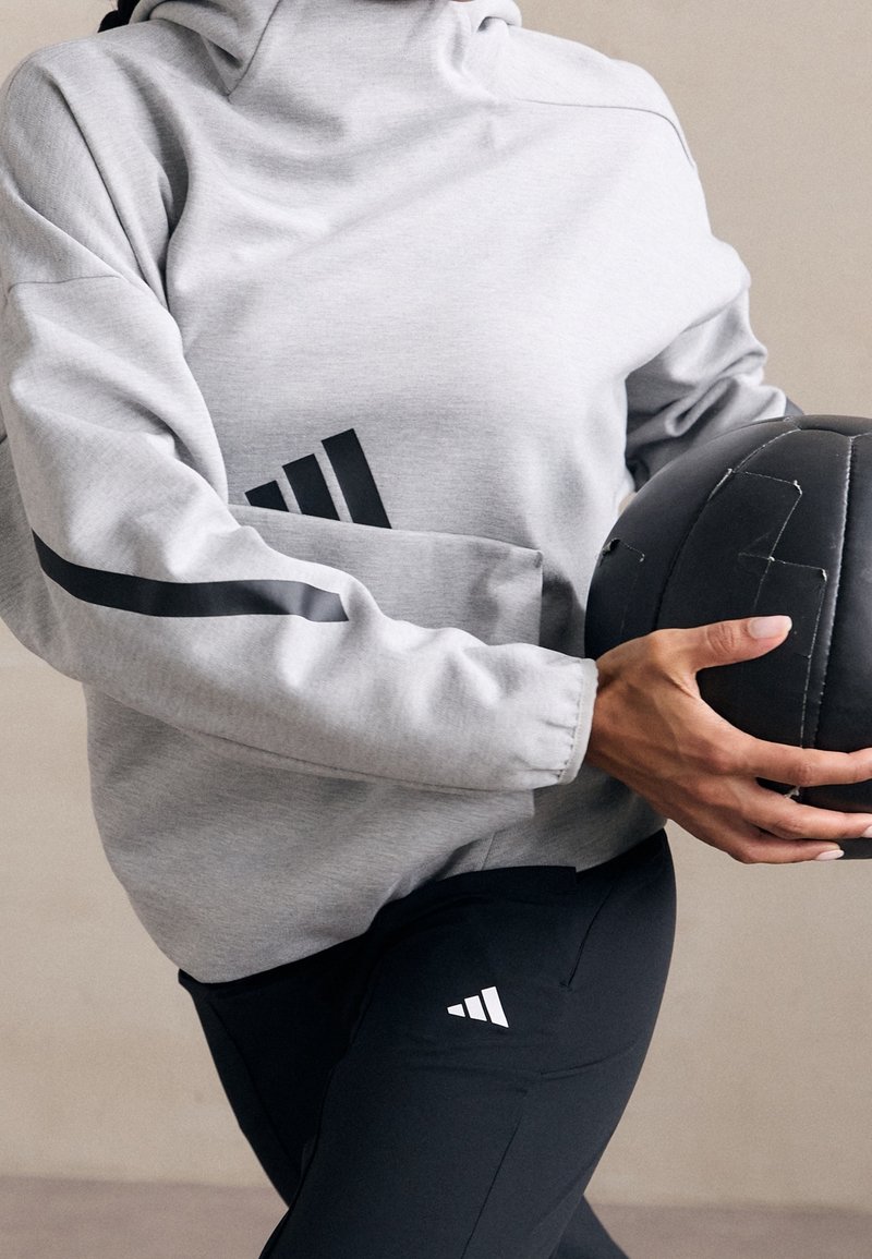 Personne tenant un ballon de médecine noir portant un sweat Adidas gris et un pantalon Adidas noir, montrant le milieu du corps et partiellement les bras.