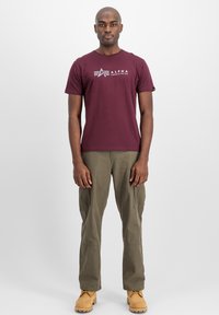 Mørkerød kortærmet t-shirt med hvid "Alpha Industries" logo; parret med olivengrønne cargo bukser og brune støvler, stående mod en ensfarvet baggrund.
