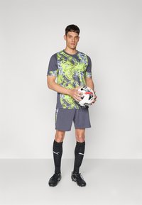 Γκρι αθλητικό μπλουζάκι με πράσινο αφηρημένο σχέδιο, ταιριαστά σορτσάκια, μαύρες ψηλές κάλτσες μέχρι το γόνατο και μαύρα ποδοσφαιρικά παπούτσια, κρατώντας μια μπάλα ποδοσφαίρου.