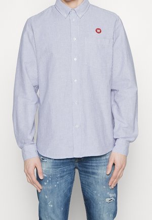 Man wearing a blue and white thin striped button-up shirt with a red circular logo on the chest pocket and distressed blue jeans.