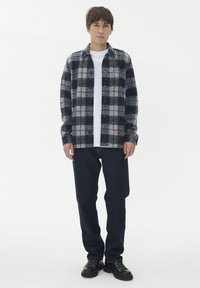 Mladenič stoji, oblečen v modro-sivo karirasto srajco s pomočnimi gumbi čez belo majico, temne kavbojke in črne vezne čevlje na beli podlagi.