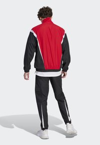 Giacca da track rossa e nera con accenti bianchi, dotata di un colletto alto e polsini elasticizzati. Abbinata a pantaloni neri, entrambi dal design sportivo.