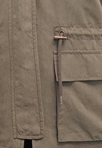 Detailní záběr na khaki bundu s texturovanou tkaninou a popruhem s kovovým přepínačem. Obsahuje náprsní kapsu se šitými akcenty.
