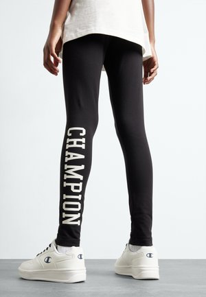 Leggings neri con un design aderente, caratterizzati da grandi scritte bianche "CHAMPION" lungo una gamba. Abbinati a sneakers bianche. Tessuto liscio e elastico.