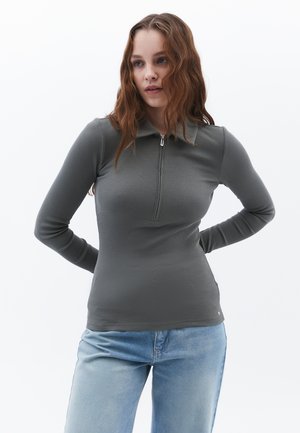 Langärmliges, graues Rippenoberteil mit Kragen und halbem Reißverschluss. Es wird mit hellblauer Jeans getragen und zeichnet sich durch eine enganliegende Passform und eine glatte Textur aus.