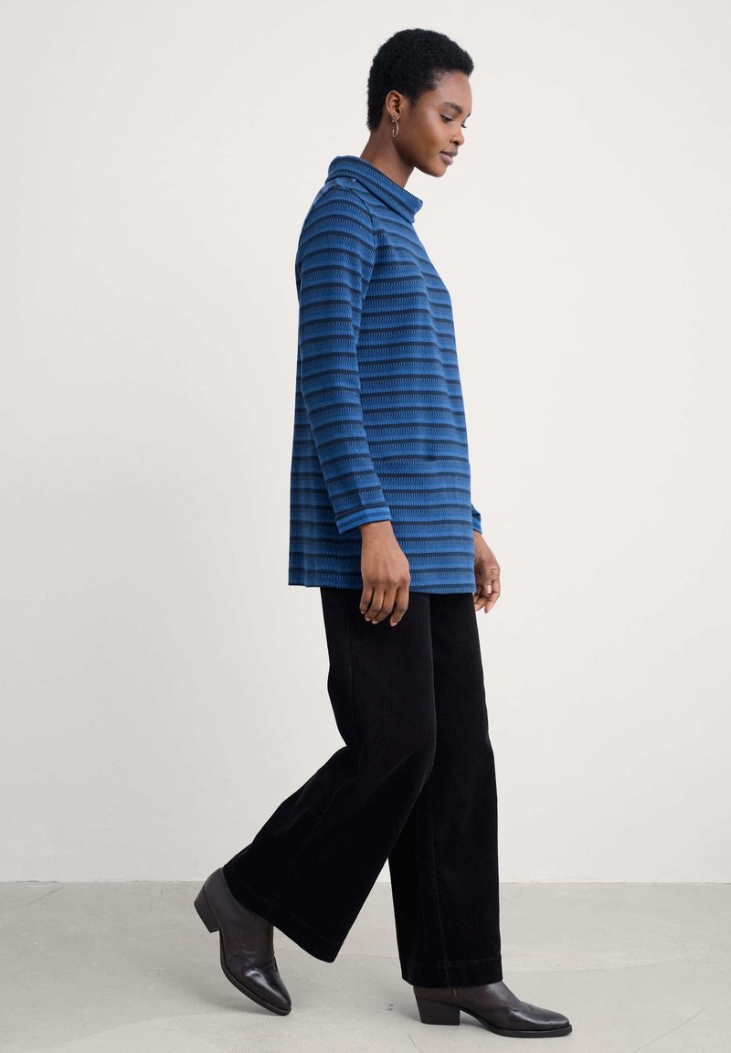 Osoba idąca w niebieskiej, pasiastej bluzce z długim rękawem, czarnych szerokich spodniach i ciemnobrązowych botkach za kostkę na jasnym tle.