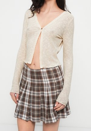 Woman wearing beige long-sleeve cropped top with one button fastened and brown plaid pleated skirt against plain background.