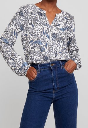Kvinde iført en hvid bluse med lange ærmer med blå paisley-mønstre stoppet ned i højtaljede mørkeblå jeans, hænderne i lommerne.