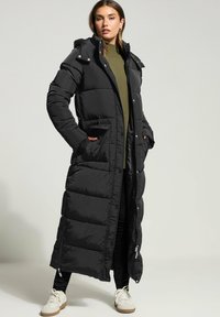 Svart lång pufferjacka med vadderad design, inklusive huva, fickor på sidorna och dragkedjeventiler. Bärs över en grön tröja och svarta byxor.