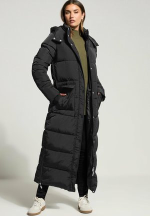 Svart lång pufferjacka med vadderad design, inklusive huva, fickor på sidorna och dragkedjeventiler. Bärs över en grön tröja och svarta byxor.