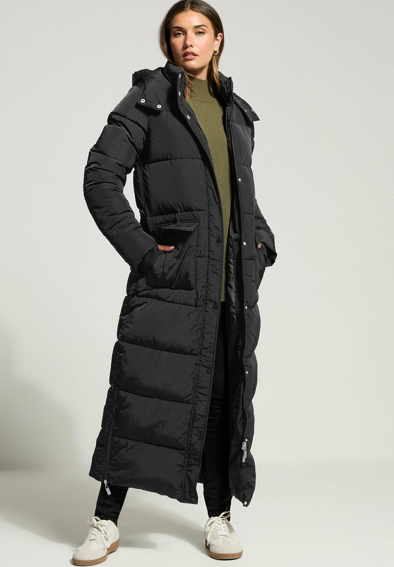 Svart lång pufferjacka med vadderad design, inklusive huva, fickor på sidorna och dragkedjeventiler. Bärs över en grön tröja och svarta byxor.