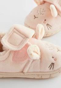 Chaussons en peluche roses avec un design de lapin, présentant des oreilles, une sangle en velcro et des détails faciaux brodés. Texture douce et semelle extérieure à motifs.