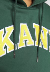 Grön hoodie med stora, broderade gula bokstäver som stavar "KANI", med vita detaljer och draremmar med metalldetaljer.