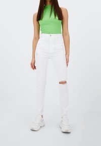 Jean blanc taille haute avec des déchirures aux genoux, associé à un débardeur ajusté en côtes vert et à des baskets blanches épaisses.