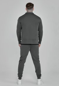Gray tracksuit with long-sleeve top featuring a collar and matching pants. Fabric appears smooth; model facing away, shoes are white sneakers.
