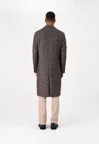 Abrigo largo en un patrón de pata de gallo en marrón y crema, confeccionado en un tejido texturizado, con una espalda con corte y un ajuste entallado. Combinado con pantalones de color claro.