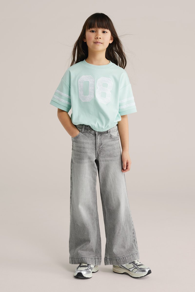 Ung pige med langt hår iført en mintgrøn skjorte med nummer 08, brede grå jeans og hvide sneakers, stående med den ene hånd i lommen.