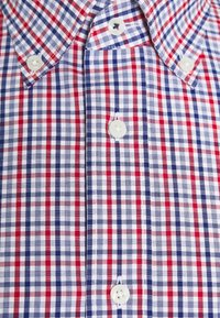 Acercamiento a una camisa de cuadros en rojo, blanco y azul, con botones blancos y cuello abotonado.