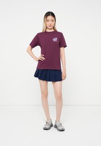 Žena stojící s rukou na boku, oblečená v fialovém tričku s grafikou, námořnické skládací mini sukni a šedých slip-on botách na bílém pozadí.
