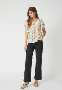 Blusa beige chiaro con pois neri, maniche corte a volant e scollatura a V. Abbinata a jeans neri a vita alta e tacchi a blocco verde opaco.