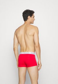 Rote Baumwollslips im Trunk-Stil mit einem weißen elastischen Taillengummi, der Branding zeigt. Das Modell ist von hinten zu sehen, der Oberkörper ist sichtbar.