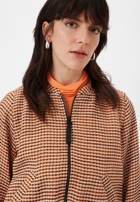 Ritssluitingjack in een oranje en wit geometrisch patroon, met een hoge kraag, gestructureerde stof en zwarte ritsdetails.