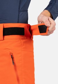 Helle orange Hosen mit einem Klettverschlussbund, ausgestattet mit einer seitlichen Reißverschlusstasche und einem eleganten Design, hergestellt aus einem strukturierten, robusten Material.