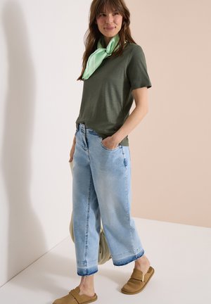 Mujer con camiseta verde oliva, vaqueros de pierna ancha azul claro con bajos deshilachados, zapatos tipo mocasín marrón claro y pañuelo verde claro al cuello, de pie con la mano en el bolsillo.