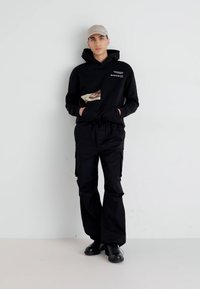 Sudadera negra con un detalle gráfico, combinada con pantalones cargo negros. El atuendo tiene un corte holgado y se complementa con una gorra ligera y botas.
