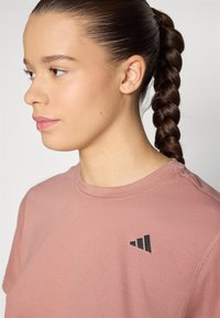 Camiseta de manga corta rosa claro hecha de tela suave con un logo negro en el pecho. Cuello redondo con un detalle de costura mínimo.