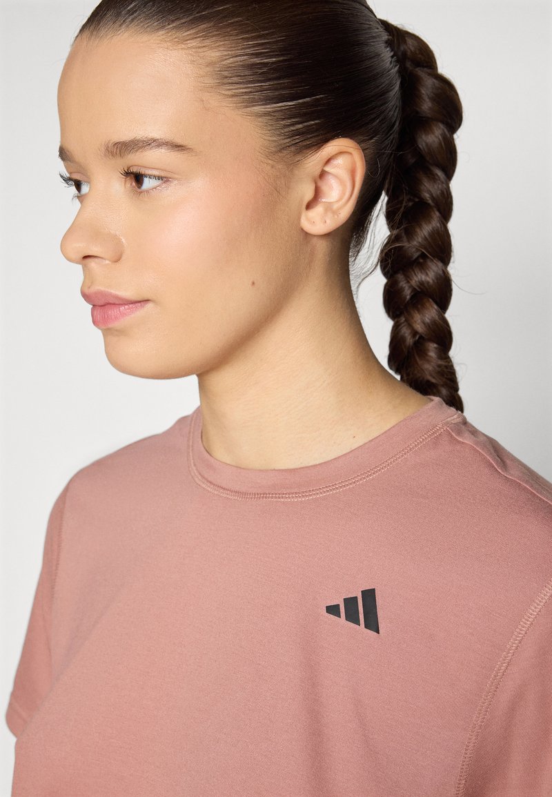 Camiseta de manga corta rosa claro hecha de tela suave con un logo negro en el pecho. Cuello redondo con un detalle de costura mínimo.