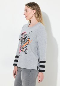 Grijze langmouwen shirt met een vissenprint in zwart, oranje en roze. Zwart-witte gestreepte accenten op de mouwen; zijsplitsingen aan de zoom.