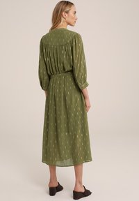 Rochie midi verde măslin cu spate plisat, mâneci trei sferturi și model cu dungi metalice. Include un centură asortată. Purtați cu încălțăminte maro.