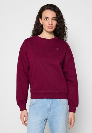 Joven mujer con cabello rizado que lleva una sudadera burdeos lisa y vaqueros azul claro, de pie frente a un fondo blanco.