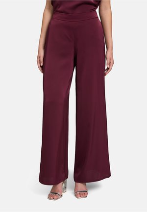 Kobieta ubrana w bordowe, szerokie spodnie z wysokim stanem, w połączeniu ze srebrnymi sandałami na klockowym obcasie z odkrytymi palcami, na jednolitym tle.