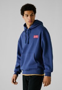 Sudadera azul marino de mezcla de algodón, con un bolsillo frontal, puños acanalados y un logotipo rojo bordado en el pecho. Diseño tipo pulóver.
