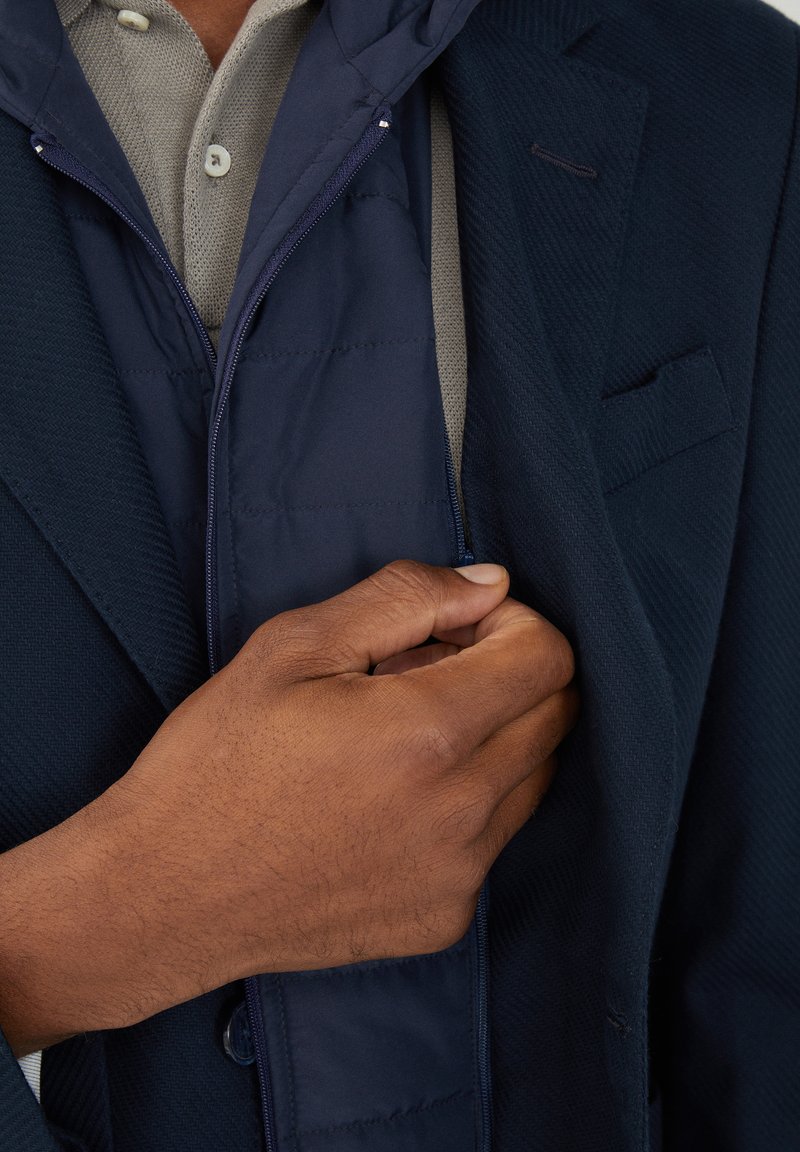La chaqueta híbrida de color azul marino presenta un forro acolchado, cierre por cremallera y capucha, superpuesta a una camisa de collar gris claro.