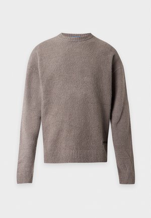 Jeune homme debout, mains dans les poches, portant un pull gris, une chemise blanche, un pantalon noir large et des baskets blanches sur fond blanc uni.