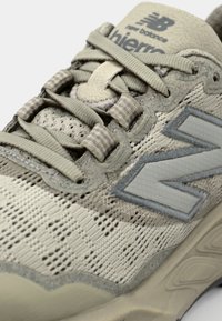 Chaussure de sport beige avec une tige en mesh, bout arrondi et coutures renforcées. Présente des accents gris texturés et un grand logo "N".
