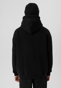 Zwarte oversized hoodie met een grote capuchon aan de achterkant, geribbelde mouwen en zoom, gecombineerd met een zwarte gebreide beanie, gepresenteerd vanuit de achterkant.