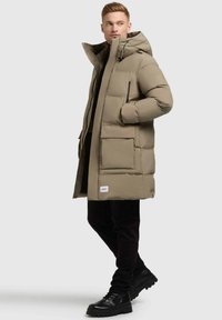 Parka beige imbottita con cappuccio, dotata di zip frontale, grandi tasche e un patch con logo. Indossata sopra un abbigliamento nero e stivali neri voluminosi.