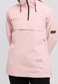 Giacca anorak rosa chiaro con tasca frontale, cerniera nera, cappuccio regolabile e patch con logo sulla manica. Texture liscia.