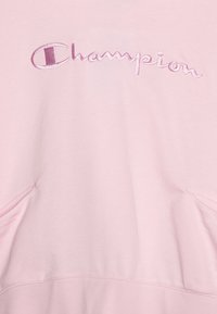 Svetloružová mikina s kapucňou vyrobená z jemného materiálu, ktorá má veľké vyšívané logo "Champion" v tmavšej ružovej farbe. Obsahuje predné vrecká a má hladkú textúru.
