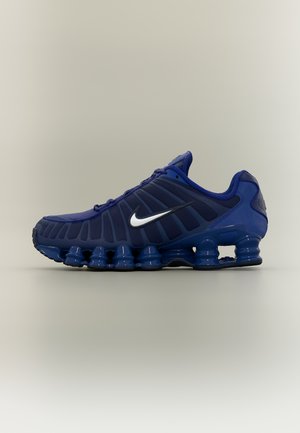 Zapatilla de running Nike Shox azul oscuro con el logo Swoosh blanco y elementos distintivos en la suela que parecen resortes, sobre un fondo neutro.
