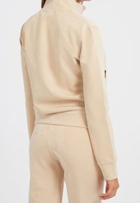 Chaqueta beige con cremallera y cuello alto, de textura suave y diseño ajustado. Presenta detalles de costura sutiles y mangas largas.